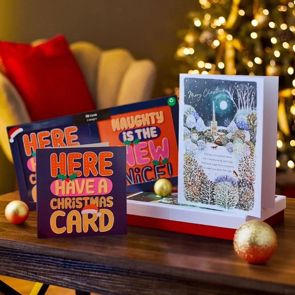 Christmas cards displayed on a table with festive decorations, including gold and red baubles, a cozy chair, and a lit Christmas tree in the background.